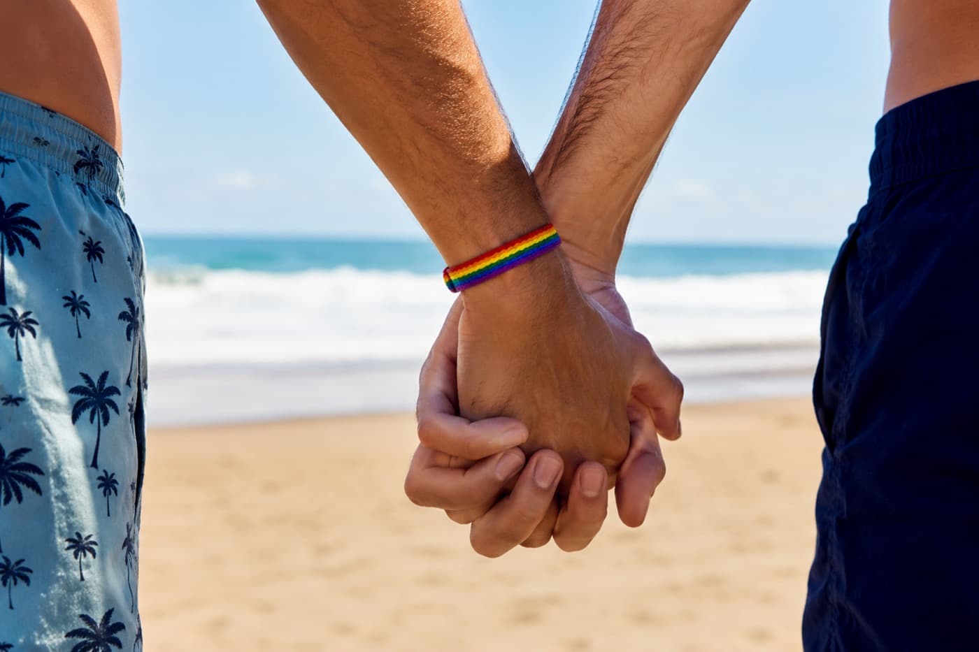 Zwei Männer halten an einem Strand Händchen, einer trägt ein Regenbogenarmband